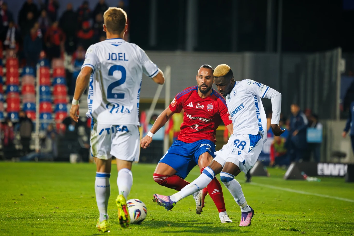 Mecz Raków Częstochowa - Lech Poznań w ramach 6. kolejki Ekstraklasy Dwaj piłkarze w białych strojach starają się zdobyć piłkę podczas meczu, podczas gdy zawodnik w czerwono-niebieskim stroju próbuje przeszkodzić w ich akcji, a w tle widoczni są kibice na trybunach.