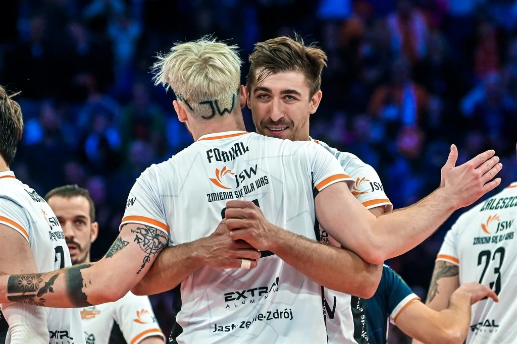 Dwóch siatkarzy drużyny w białych strojach ściska się na boisku po zakończonym meczu, wokół widać innych zawodników, a za nimi rozmyte światła i publiczność.