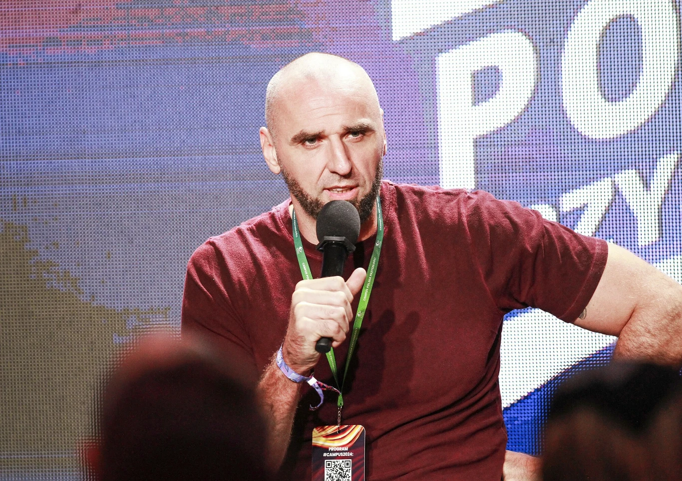 Marcin Gortat Mężczyzna z brodą i ogoloną głową w bordowej koszulce mówi do mikrofonu, siedząc na tle kolorowego baneru z napisem, w ręku ma przepustkę na smyczy.