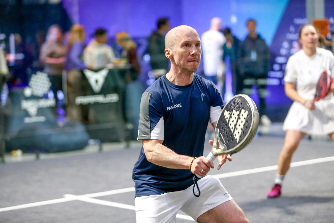 Raper Mezo grający w padla Mężczyzna w sportowym stroju trzyma rakietę do padla, koncentrując się na akcji podczas meczu, w tle widoczna druga zawodniczka oraz rozmyta publiczność.