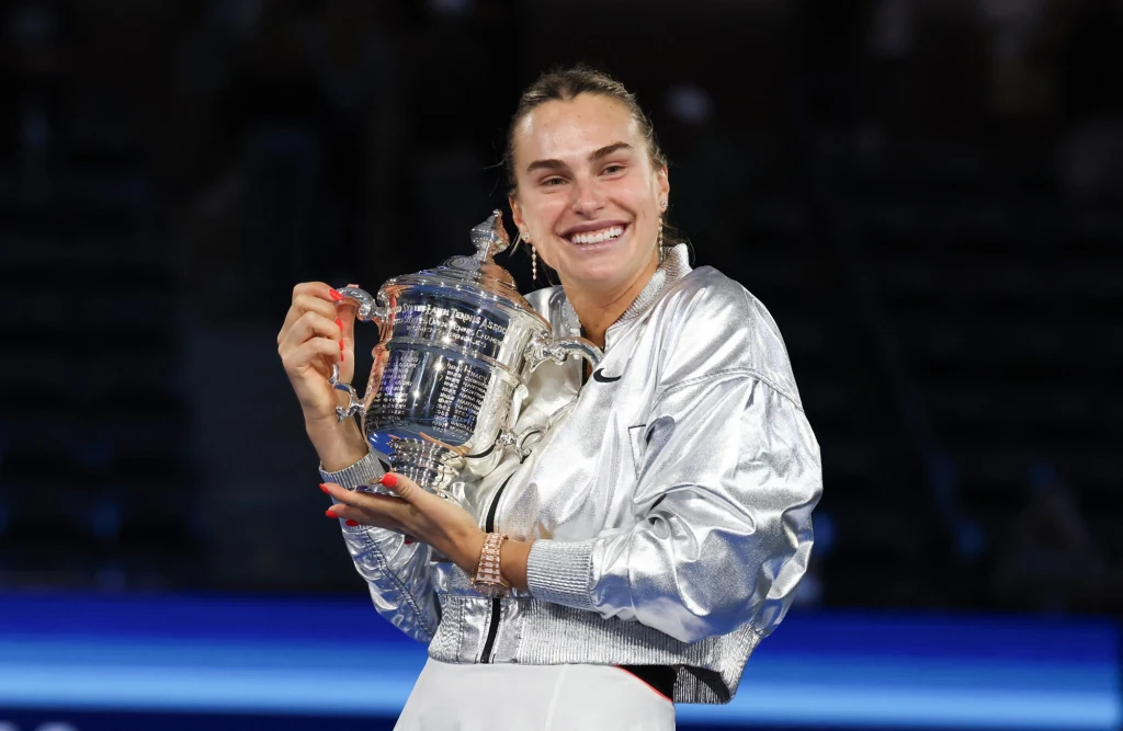Aryna Sabalenka Uśmiechnięta kobieta w srebrnej kurtce trzyma w rękach duży, ozdobny puchar, stojąc na tle rozświetlonego kortu tenisowego.