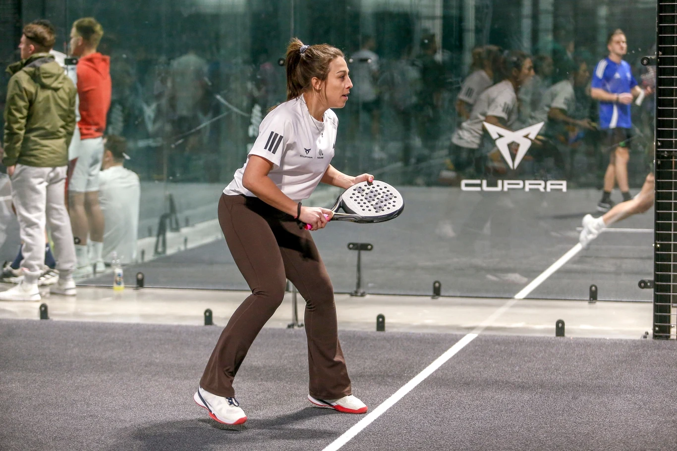 Joanna Jędrzejczyk grająca w padla Kobieta w sportowym stroju przygotowuje się do odbicia piłki na korcie do padla, w tle kilku obserwatorów oraz szklane ściany kortu.