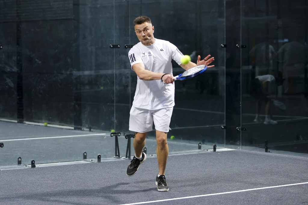 Mężczyzna w sportowym stroju odbija piłkę rakietą na korcie do padla, skupiony na dynamicznej akcji podczas gry.