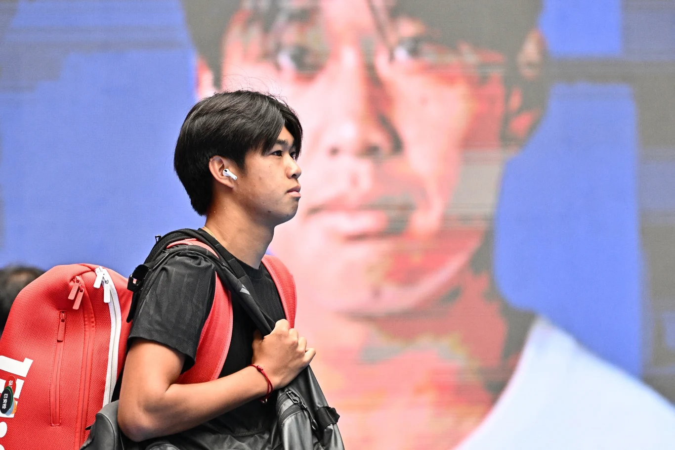 Learner Tien Młody mężczyzna z czarnymi włosami idzie z torbą sportową na ramieniu, w tle duży ekran z rozmytym wizerunkiem jego twarzy w dużym powiększeniu.