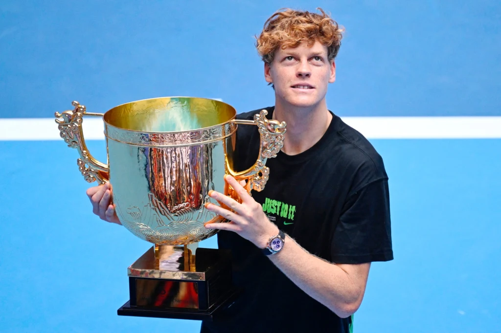 Młody mężczyzna z rudymi włosami w czarnym t-shircie trzyma duży, złoty puchar z ornamentami na tle niebieskiego kortu tenisowego, patrzy lekko w górę z uśmiechem triumfu.
