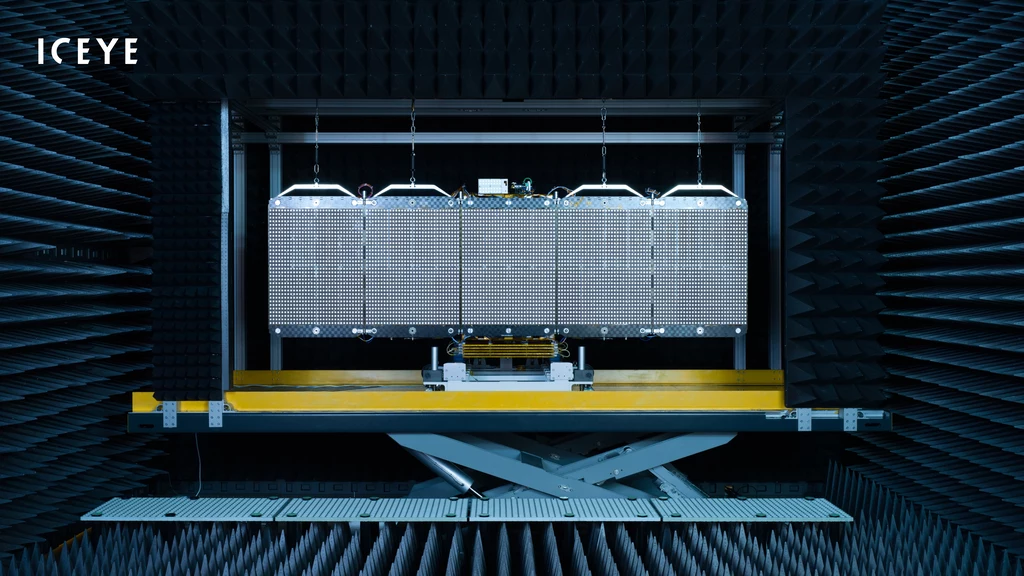 Antenna satelitarna ICEYE umieszczona w komorze bezechowej do testów technologicznych, otoczona pianką tłumiącą fale radiowe oraz oświetlona lampami technicznymi.
