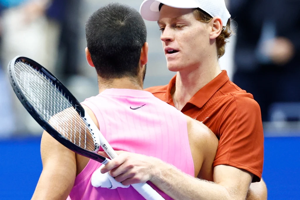 Dwóch tenisistów wymienia uścisk po zakończonym meczu, jeden ubrany w różową koszulkę, drugi w czerwoną z białą czapką, w tle nieostre trybuny.