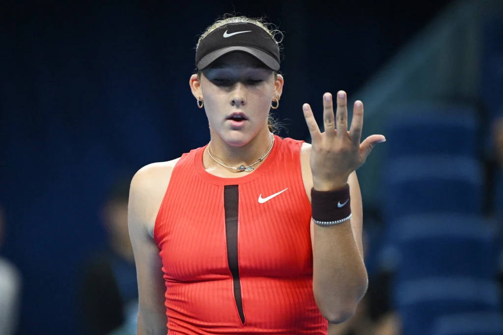 Tenisistka ubrana w czerwony strój sportowy i czarną czapkę z daszkiem, trzymająca rękę przed sobą i patrząca na nią podczas meczu na korcie tenisowym.