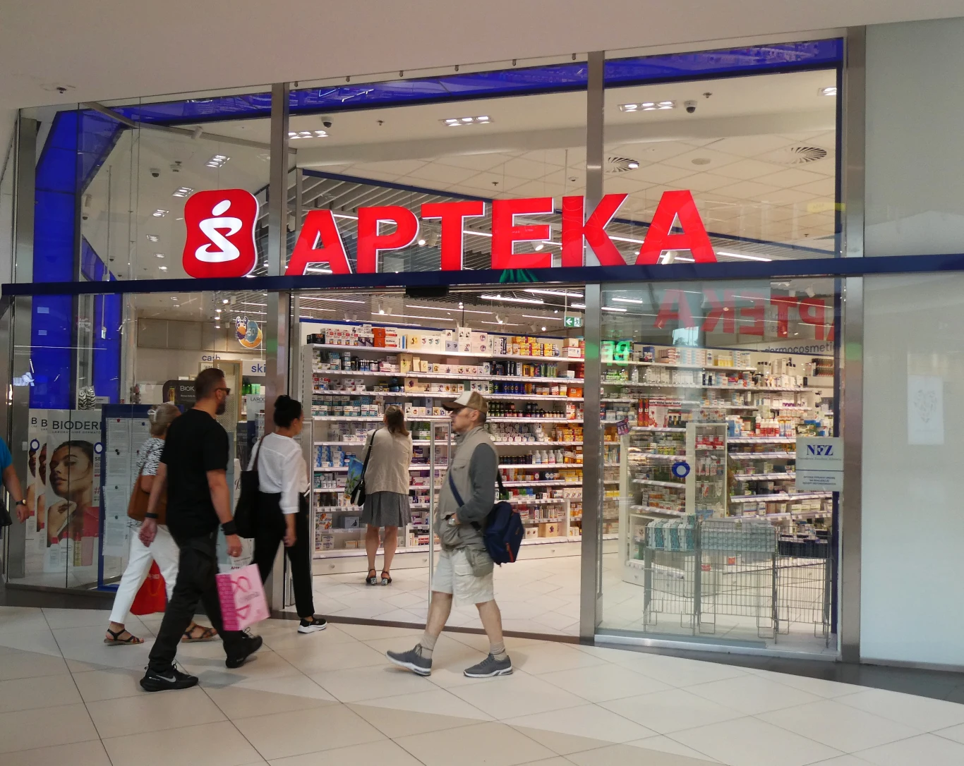 Apteki będą musiały przyjmować wszystkie butelki i puszki? Właściciele chcą zmiany przepisów. Wejście do nowoczesnej apteki w centrum handlowym z przeszklonym frontem, czerwonym napisem 'APTEKA' i przechodniami wchodzącymi do środka, witryna prezentuje półki z lekami i artykułami medycznymi.