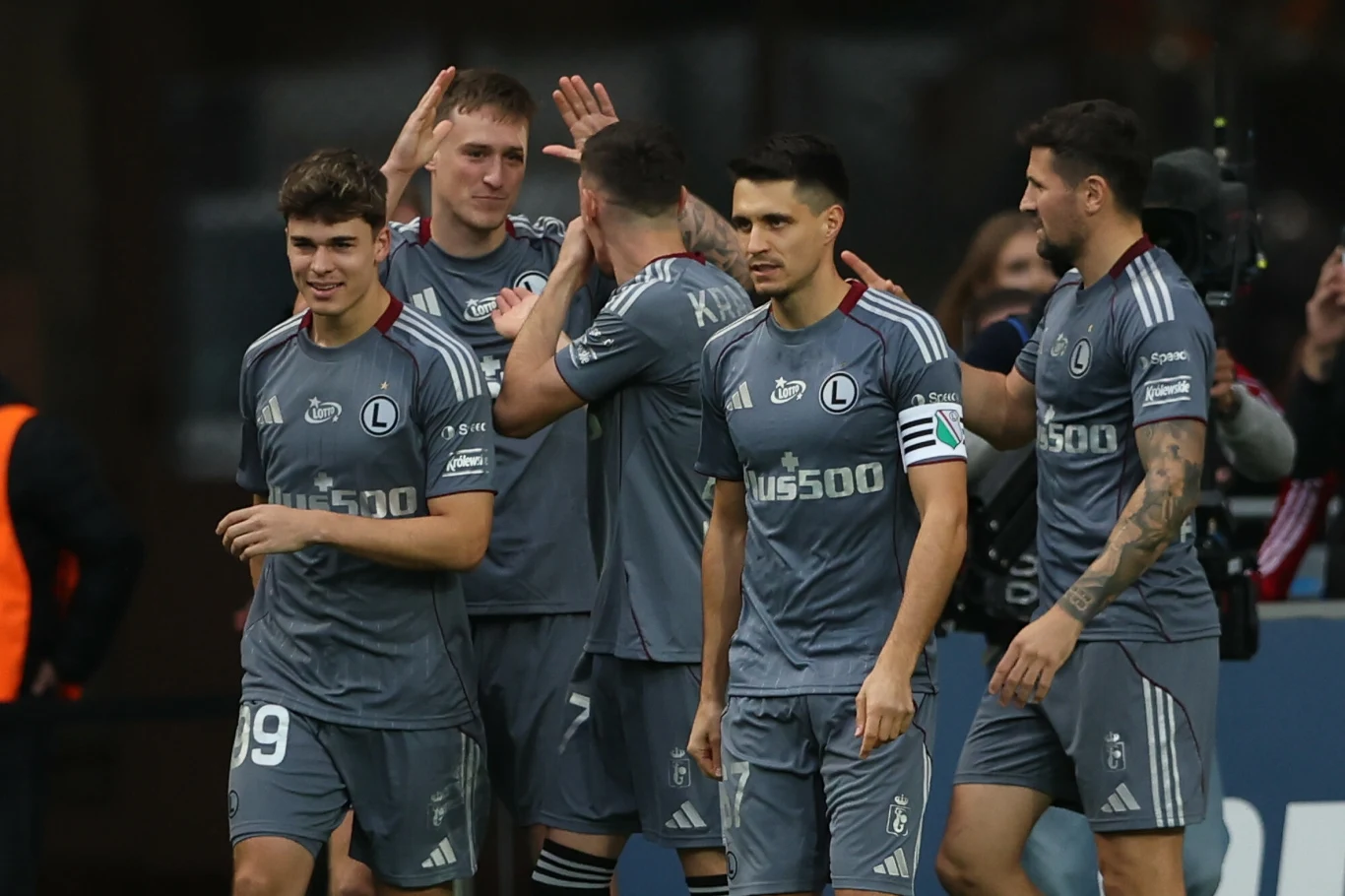 Legia Warszawa Grupa piłkarzy w szarych strojach sportowych świętuje bramkę lub ważny moment podczas meczu, widoczny uśmiech na twarzach, gesty uznania oraz logo sponsora na koszulkach