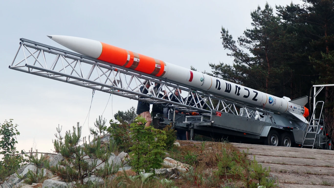 Perun gotowy do startu. SpaceForest zbuduje jeszcze większą rakietę. Rakieta badawcza w pomarańczowo-białych barwach umieszczona na platformie startowej w otoczeniu leśnym, przygotowywana przez grupę osób do wystrzelenia.