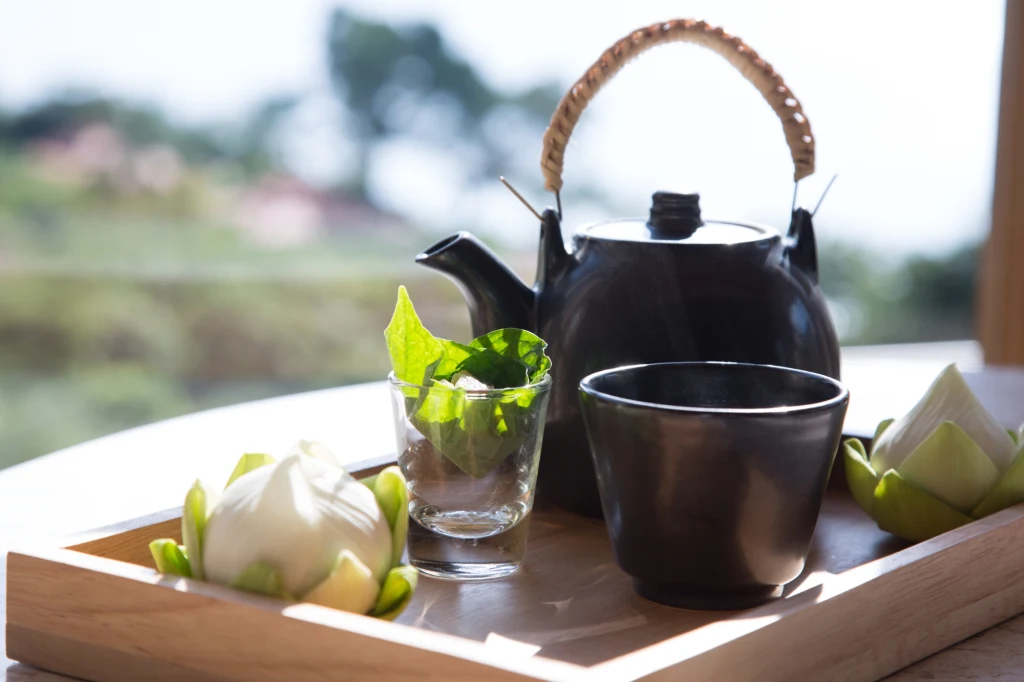 Czarna ceramiczna czajniczka z bambusowym uchwytem ustawiona na drewnianej tacy obok filiżanki, szklanki z liściem oraz dwoma kwiatami lotosu.