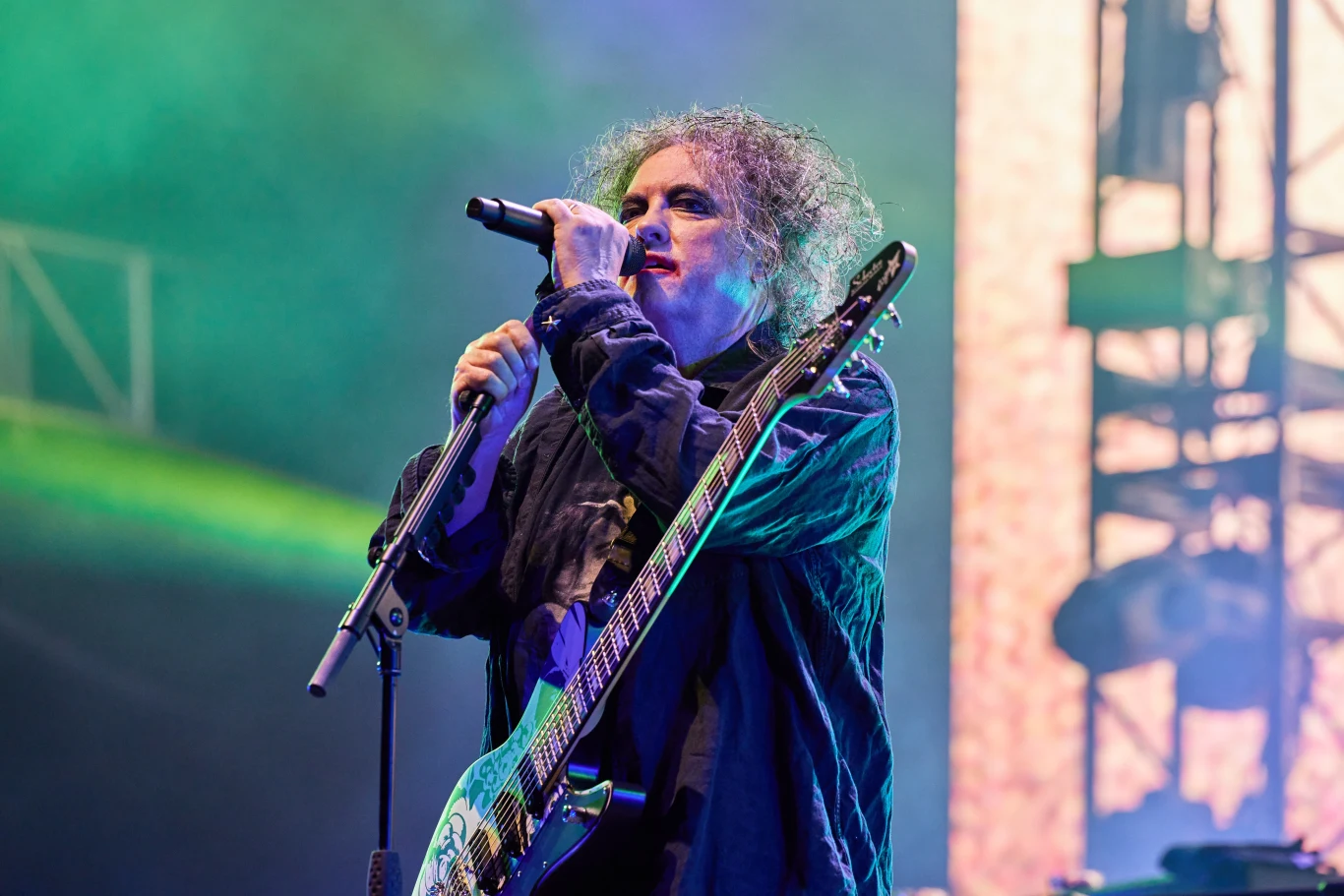 Robert Smith stoi na czele The Cure Mężczyzna o charakterystycznej fryzurze śpiewający do mikrofonu podczas koncertu, trzymający gitarę elektryczną, kolorowe światła sceniczne i części konstrukcji sceny w tle.