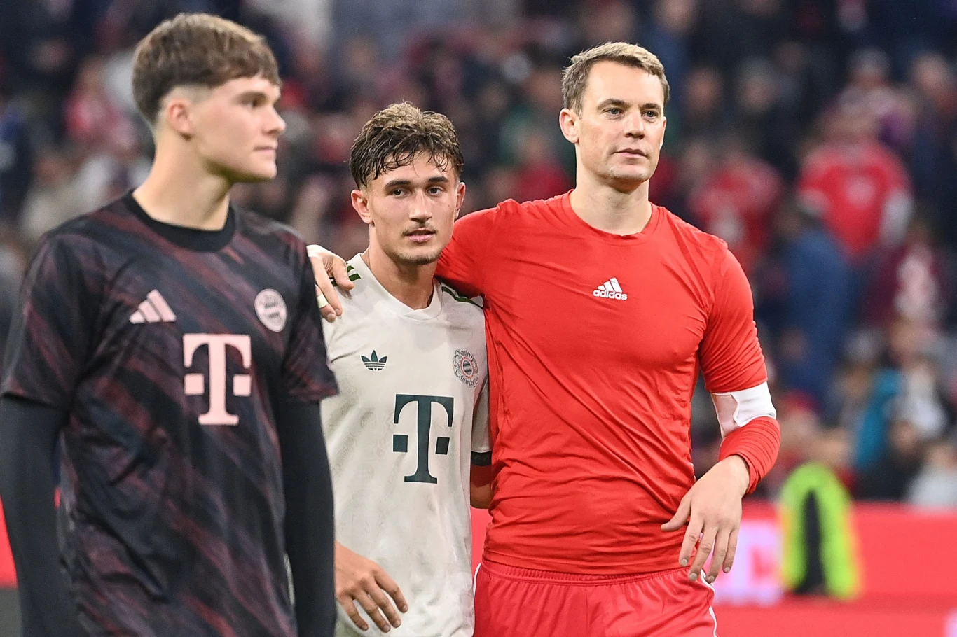 Trzech piłkarzy drużyny Bayern Monachium w strojach sportowych, jeden z zawodników obejmuje drugiego ramieniem, w tle rozmazany tłum kibiców na stadionie.