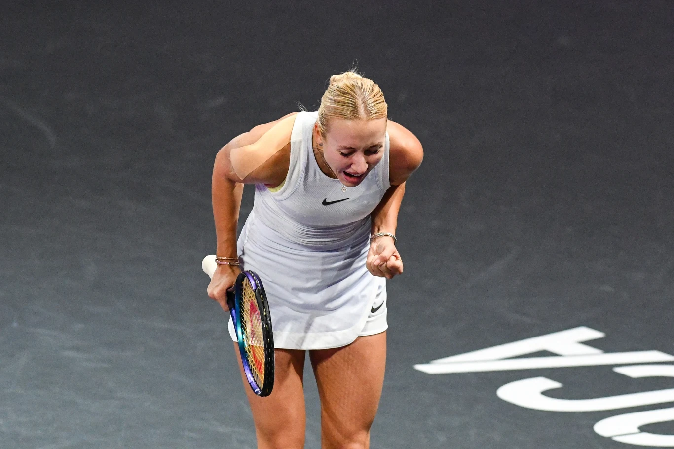 Anastazja Potapowa Zawodniczka tenisowa w białej sportowej sukience z rakietą w dłoni wyraża silne emocje po udanym zagraniu, stojąc na ciemnym korcie.