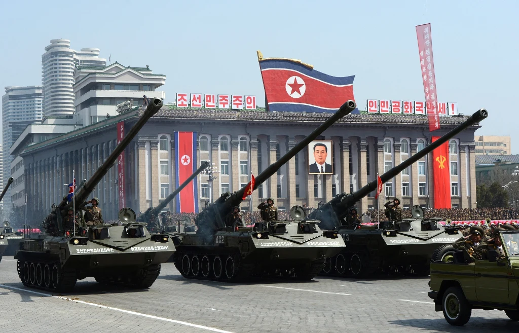 Czołgi z wysuniętymi lufami przemieszczają się w szyku na tle dużego budynku ozdobionego portretem oraz flagą Korei Północnej, wokół widać żołnierzy i elementy oficjalnej defilady wojskowej.