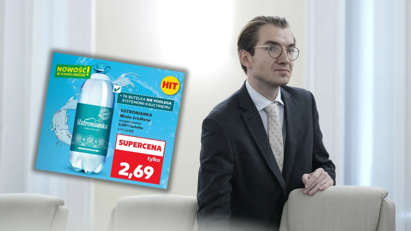 Ustronianka kontra Szyszko – spór o butelki 3,001 Mężczyzna w okularach i garniturze stoi obok białego fotela, z lewej strony widoczna jest reklama wody mineralnej z informacją o promocyjnej cenie.