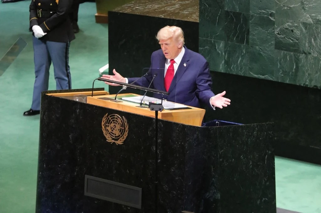 Mężczyzna w garniturze i czerwonym krawacie przemawia z mównicy oznaczonej symbolem Organizacji Narodów Zjednoczonych, w tle fragment marmurowej ściany, obok stoi umundurowany strażnik.