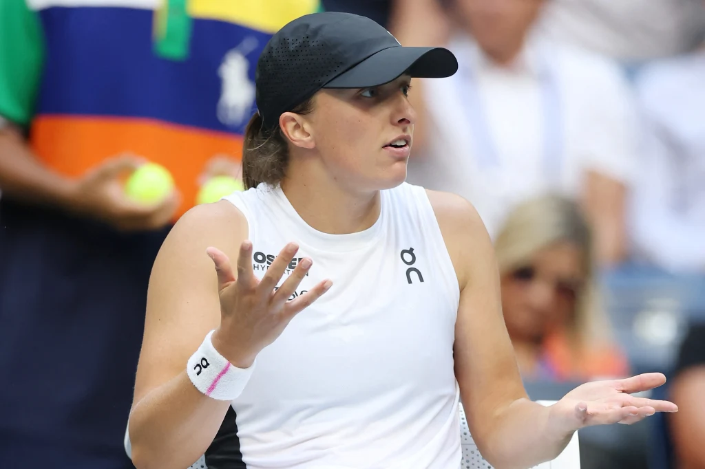 Tenisistka w białym stroju sportowym i czapce z daszkiem wykonuje gest zdziwienia lub niedowierzania, siedząc na korcie tenisowym, w tle widoczne są rozmyte postacie i fragmenty kolorowych banerów.