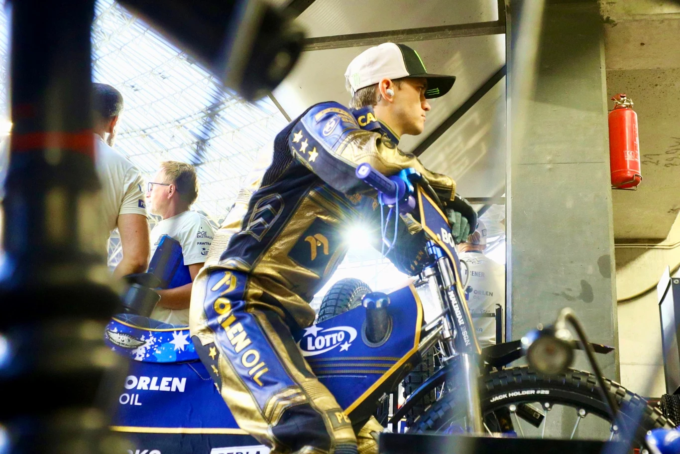 Zawodnik sportowy w stroju wyścigowym przygotowuje się do startu na motocyklu żużlowym, widoczne elementy sponsorów i sprzętu technicznego, w tle członkowie zespołu serwisowego.