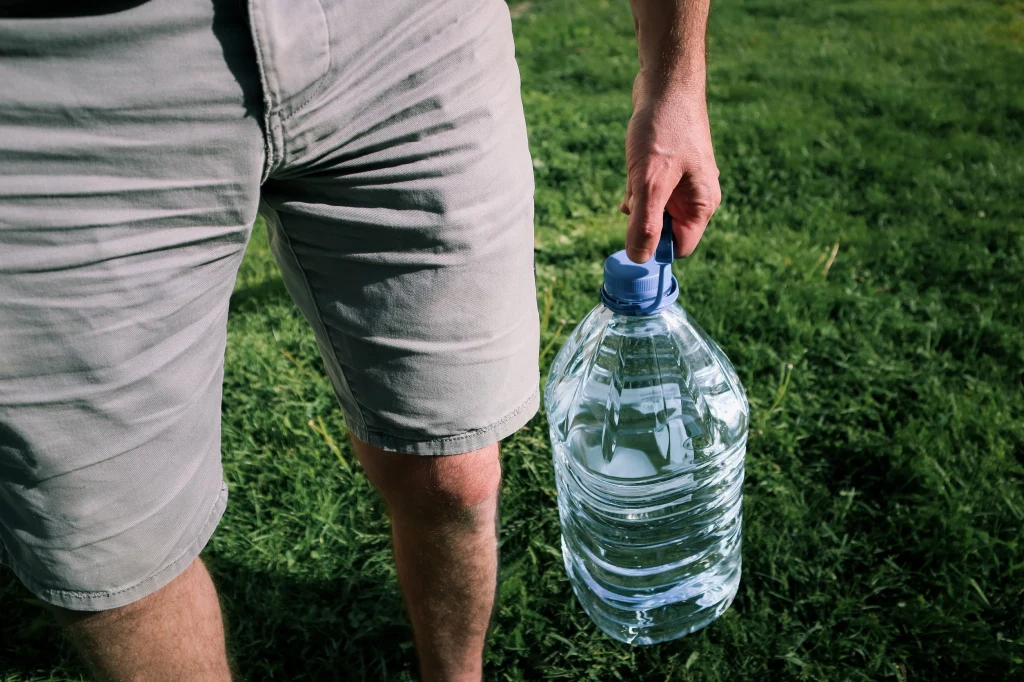 Mężczyzna w szortach trzymający dużą plastikową butlę wody, stojąc na trawie.