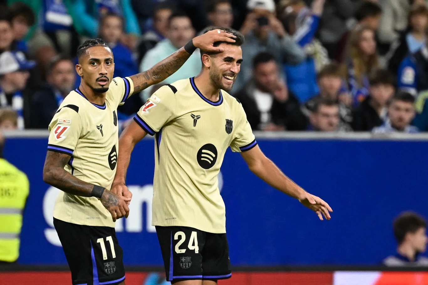 Dwóch piłkarzy drużyny FC Barcelona w jasnych strojach świętuje zdobycie gola na boisku, jeden delikatnie głaszcze drugiego po głowie, w tle widoczni kibice oraz fragment trybun.