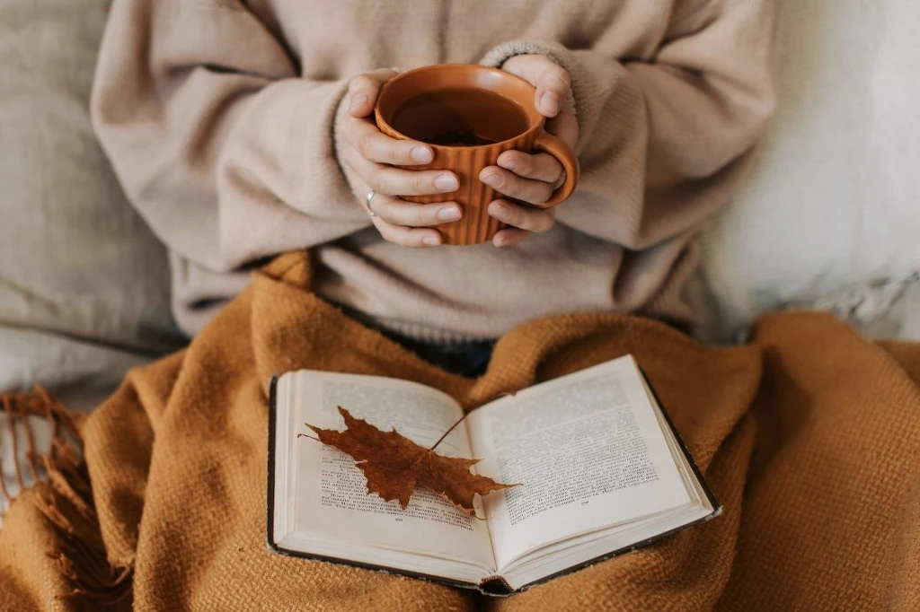 Otwarta książka leżąca na brązowym kocu, na jej stronach znajduje się suchy liść, osoba trzyma w dłoniach ceramiczny kubek z gorącym napojem.