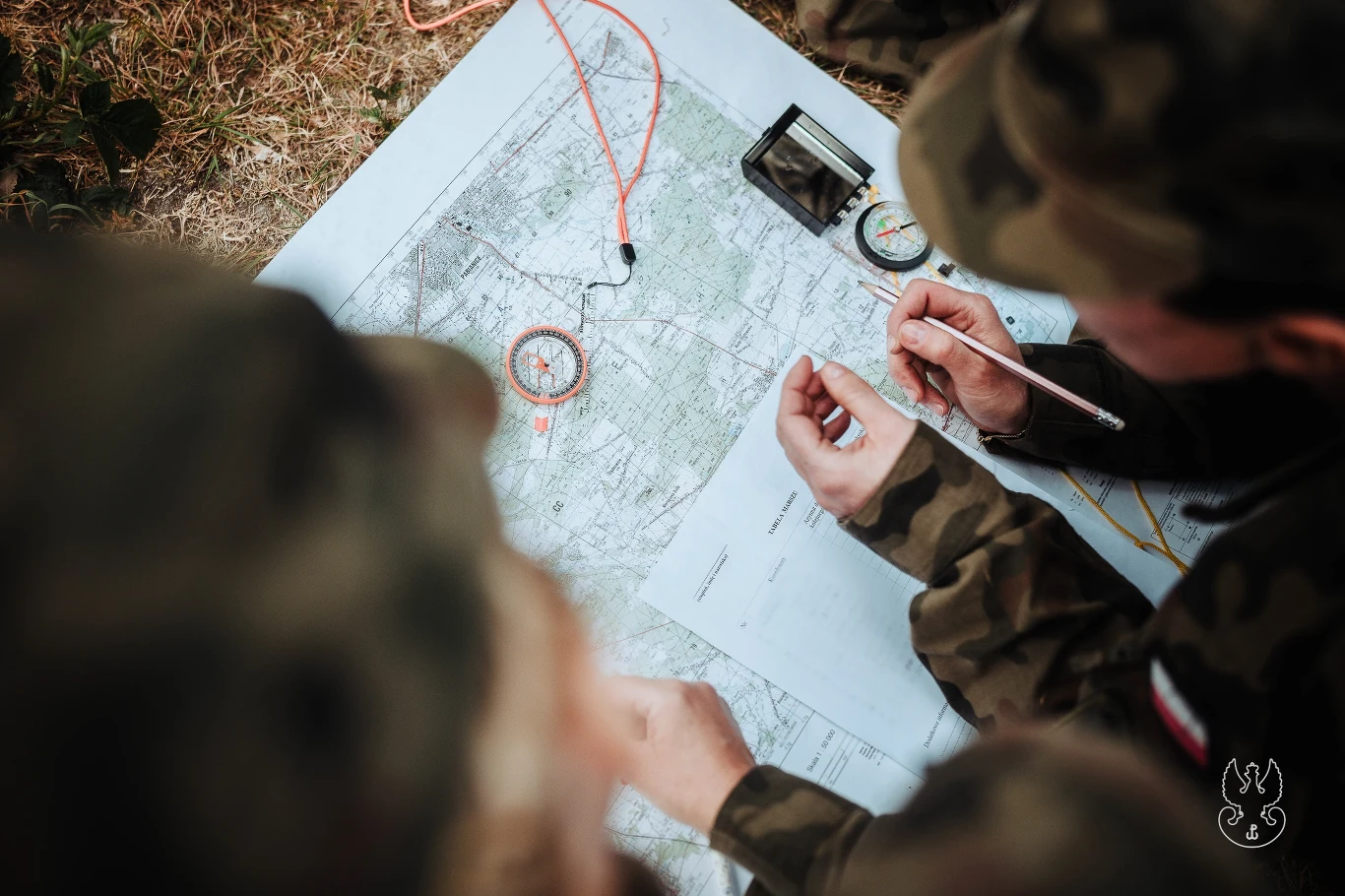 Ile zarabiają terytorialsi? Mapy topograficzna rozłożona na ziemi, wokół której siedzą osoby w mundurach wojskowych, trzymające ołówki, kompas i notatki, skupione na analizie terenu i planowaniu.