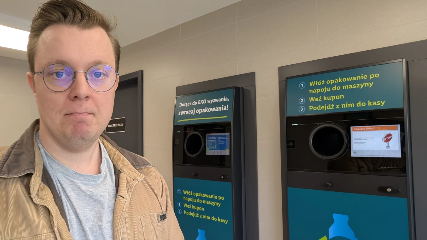 Młody mężczyzna w okularach stoi obok automatów przyjmujących butelki i puszki w pomieszczeniu o nowoczesnym wystroju, na ekranach automatów widoczne są krótkie instrukcje obsługi.