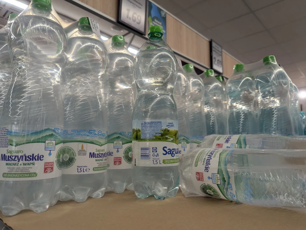 Duże ilości plastikowych butelek z wodą mineralną różnych marek ustawione są na sklepowej półce, niektóre leżą przewrócone, a całość oświetlona jest światłem sklepowym.