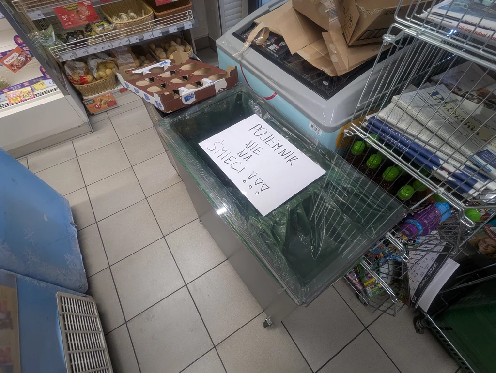 Otwarte chłodziarko-zamrażarka w sklepie spożywczym, przykryta folią, na której leży kartka z napisem „Pojemnik nie na śmieci!!!”. W tle widoczne są półki z produktami spożywczymi i kartony.