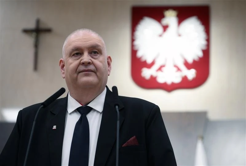 Prezes Trybunału Konstytucyjnego Bogdan Święczkowski Mężczyzna w garniturze i pod krawatem stoi za dwoma mikrofonami, w tle widoczny duży herb Polski z białym orłem oraz krzyż na ścianie.