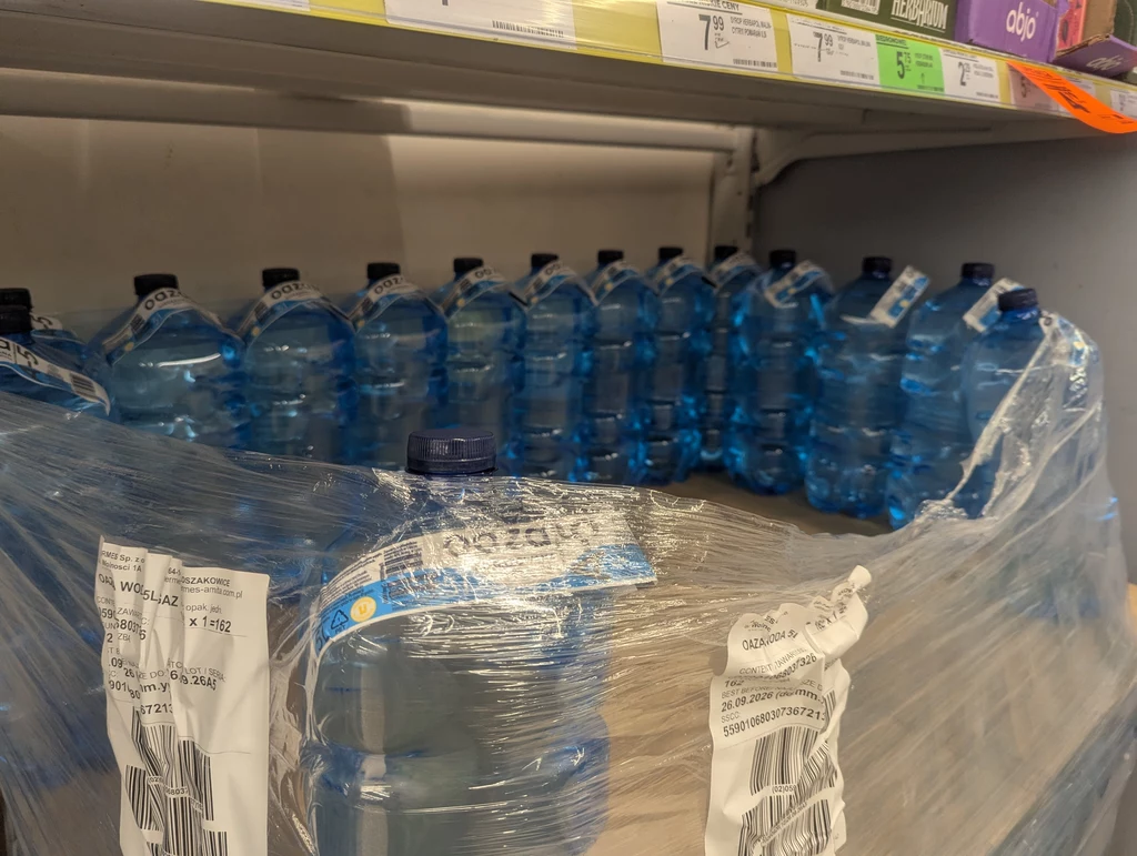 Zgrzewka plastikowych butelek wody mineralnej ustawiona na półce sklepowej, przezroczysta folia zabezpieczająca opakowanie, widoczne etykiety logistyczne na folii.
