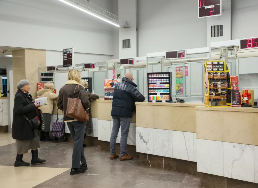 Trzy osoby stoją przy okienkach w urzędzie, część z nich obsługiwane przez pracowników za szybą, na ladzie wystawione są produkty takie jak przekąski i napoje, widoczne są także tablice z numerami do obsługi.
