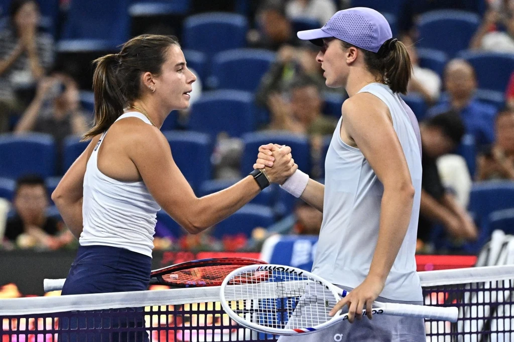 Dwie tenisistki podają sobie ręce nad siatką po zakończonym meczu na profesjonalnym korcie, wokół widownia i niebieskie trybuny.