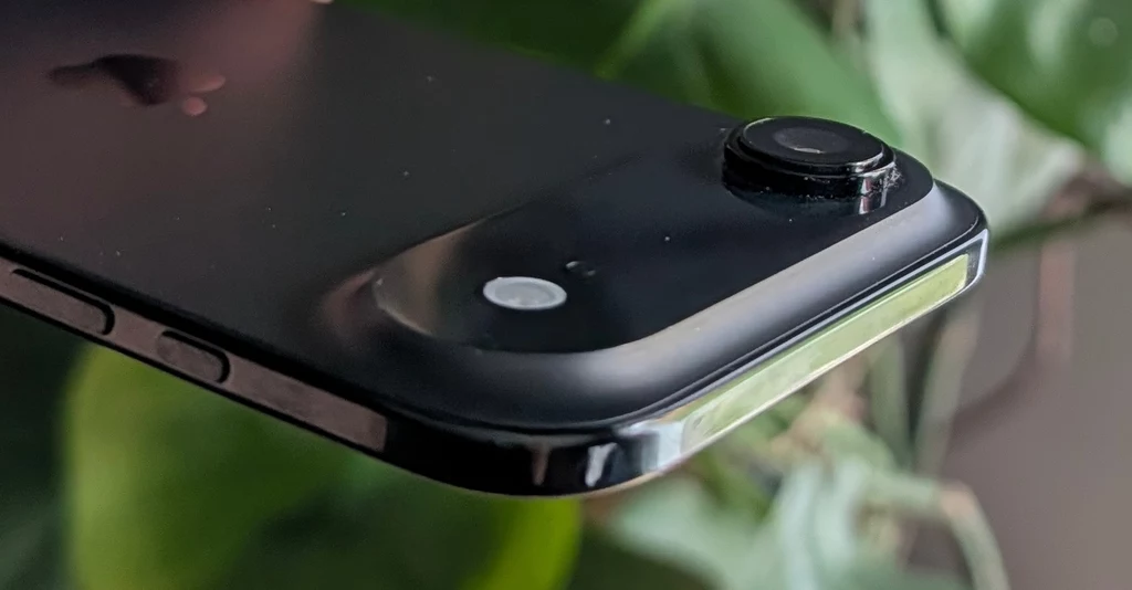 Czarny smartfon z wyraźnie widocznym obiektywem aparatu, który odstaje od powierzchni tylnej obudowy, z rozmytym zielonym tłem roślinnym.