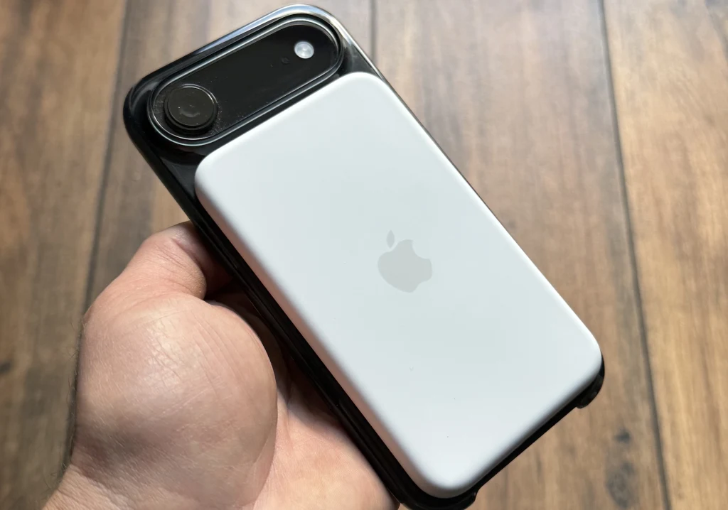 Ręka trzyma smartfon z białym akcesorium marki Apple przymocowanym na tylnej części obudowy, na tle drewnianej podłogi.