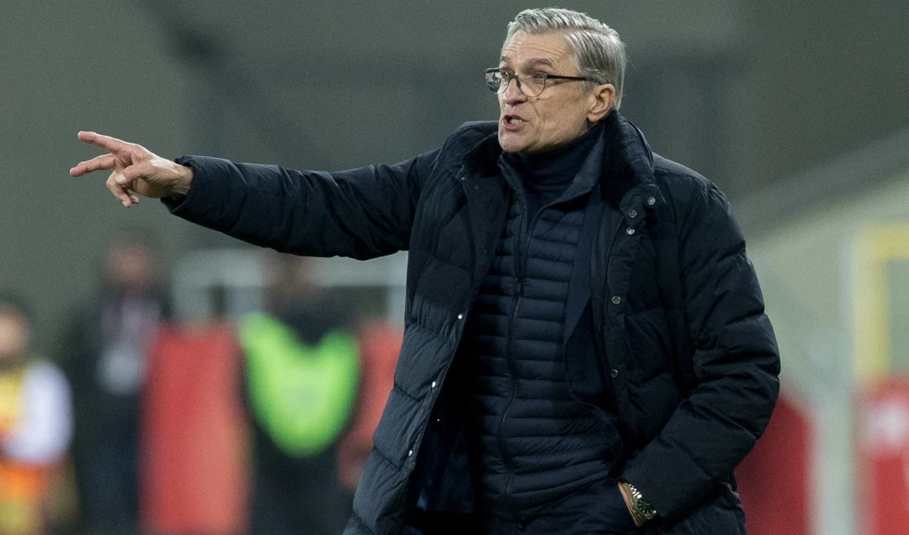 Adam Nawałka Mężczyzna w okularach i ciemnej kurtce puchowej gestykuluje ręką, wskazując coś po lewej stronie kadru, tło rozmyte z widocznymi kolorowymi elementami i osobami w tle.