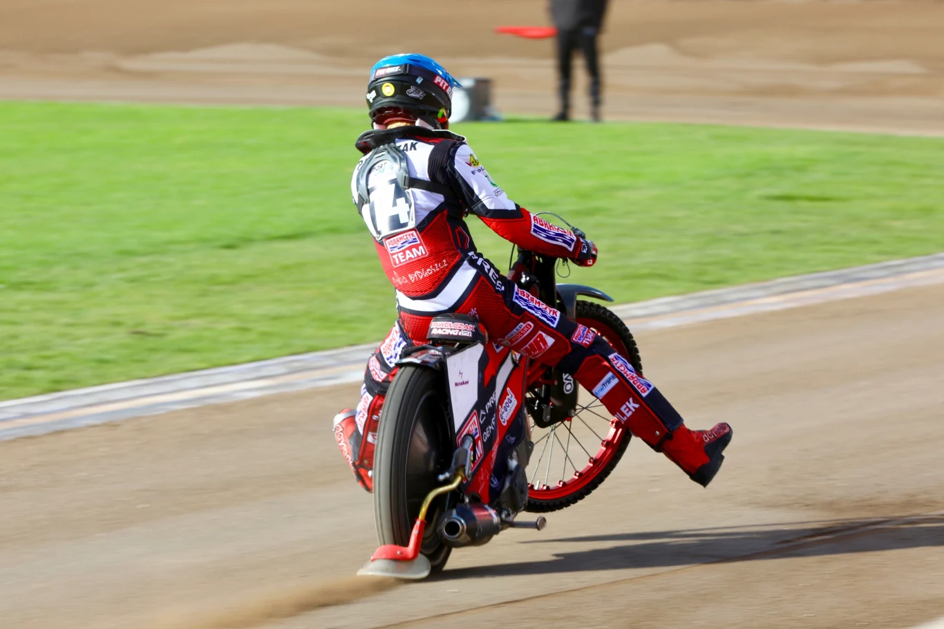 Zawodnik sportów motorowych na motocyklu wykonujący efektowny manewr na torze żużlowym, ubrany w kombinezon wyścigowy i kask, dynamiczny ruch koła oraz kurz wzbijający się spod opony.