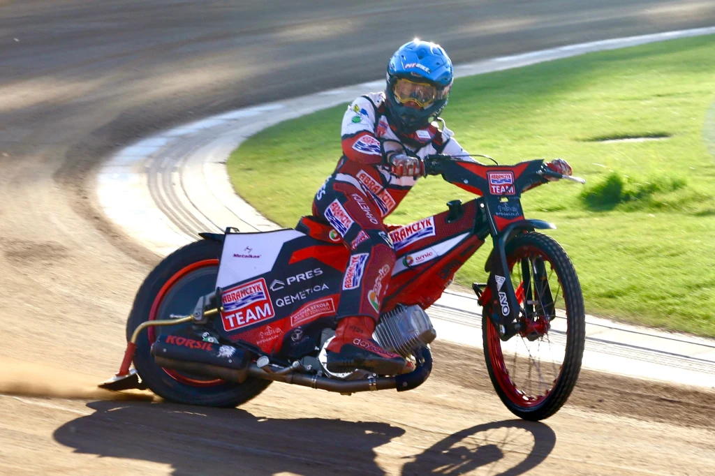 Maksymilian Pawełczak Zawodnik żużlowy w pełnym stroju ochronnym pokonuje zakręt na torze, dynamicznie pochylając motocykl; w tle wyraźnie widoczny jest fragment toru żużlowego oraz zielona murawa.