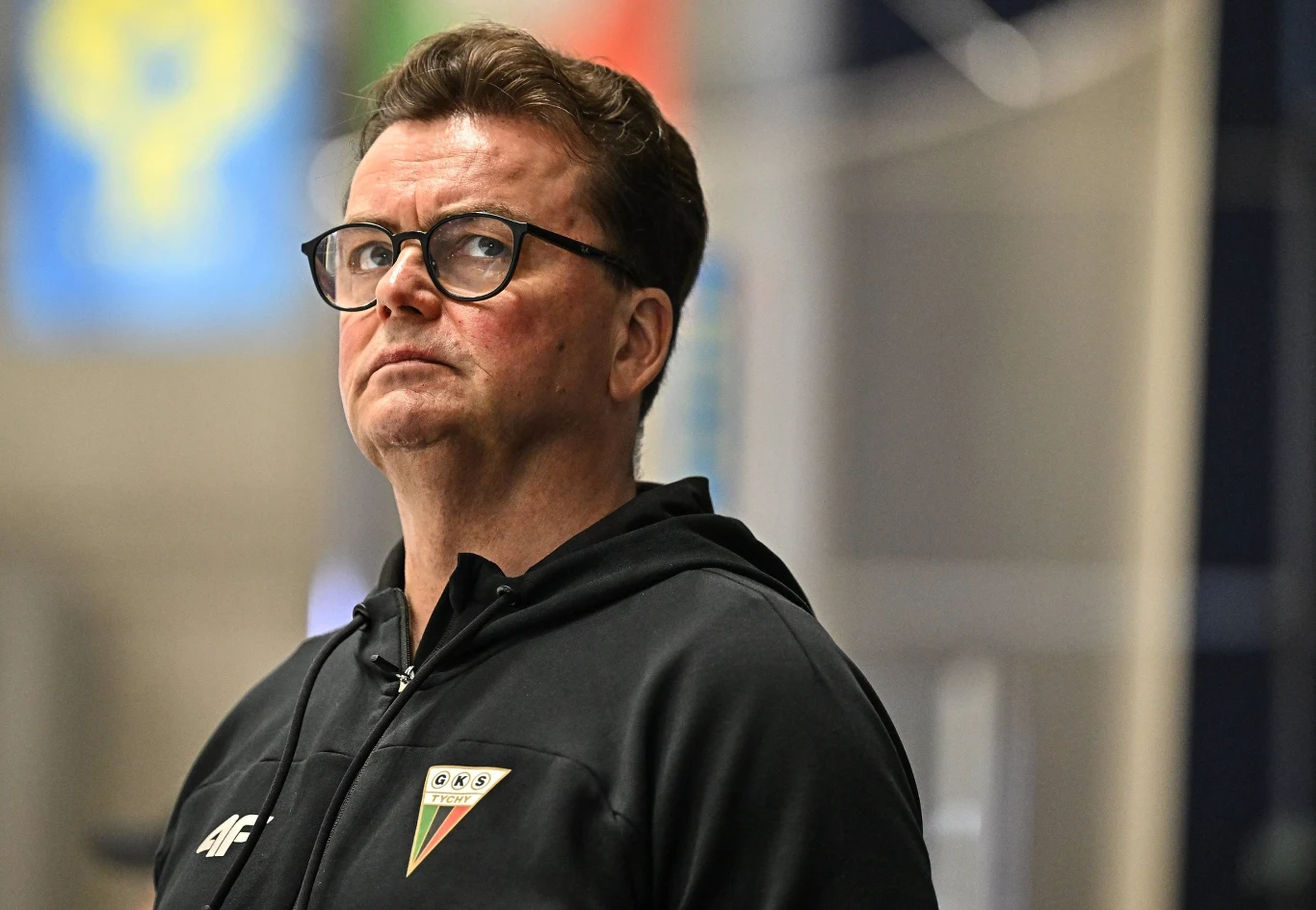 Mężczyzna w okularach ubrany w czarną bluzę sportową z logo klubu sportowego na piersi, stoi w lekkim półprofilu i patrzy w górę w skupieniu, w tle rozmyte barwne elementy.