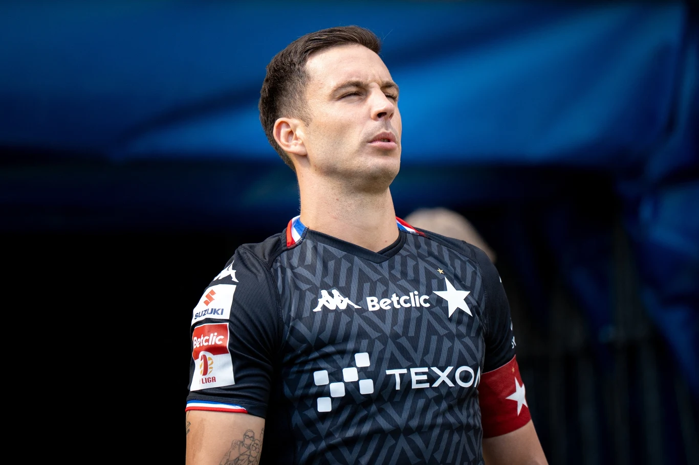 Angel Rodado Piłkarz w czarnej koszulce drużyny Wisła Kraków z widocznymi logotypami sponsorów, na tle rozmytego niebieskiego tła, skupiony i patrzący w dal.