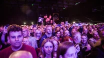 Tłum ludzi uczestniczących w wydarzeniu muzycznym, wielu trzyma telefony i robi zdjęcia lub nagrywa, w tle widoczne balony i światła sceniczne, twarze uczestników oświetlone barwnym światłem.
