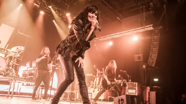 Wokalistka w ciemnych ubraniach śpiewa na scenie koncertowej przy intensywnym oświetleniu, w tle widoczni pozostali członkowie zespołu grający na instrumentach.