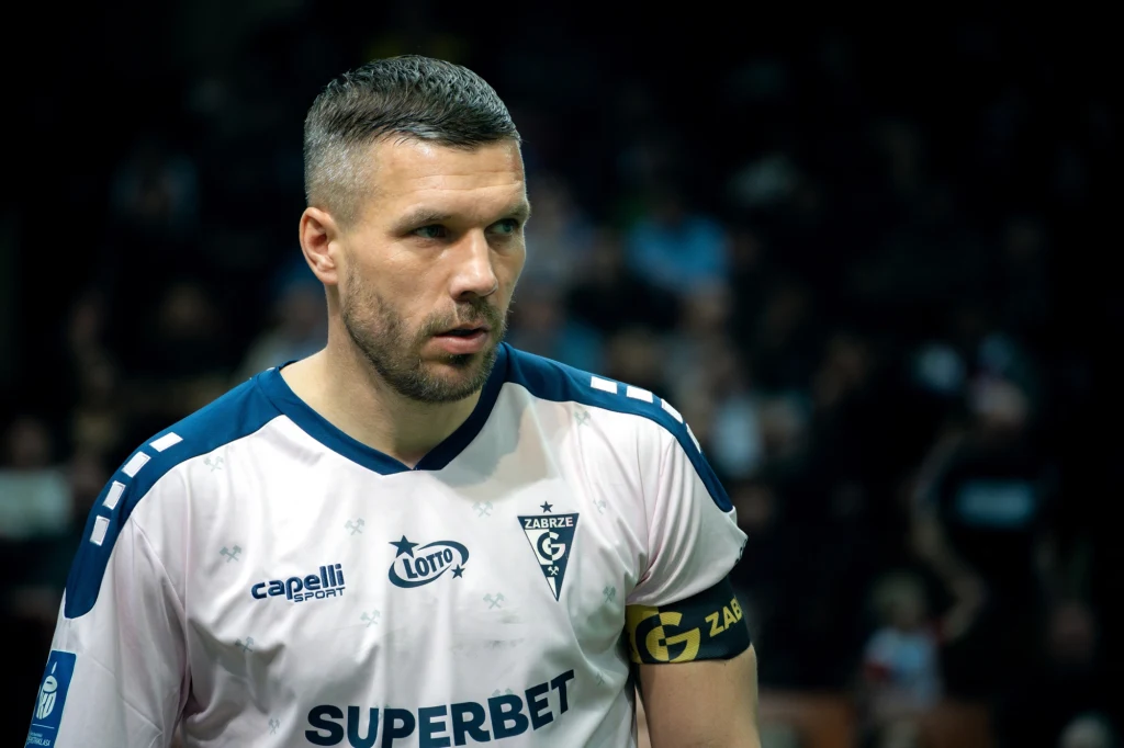 Lukas Podolski Piłkarz w biało-niebieskiej koszulce z opaską kapitana na ramieniu, widoczne logo klubu Górnik Zabrze oraz sponsorzy, w tle rozmyta publiczność.