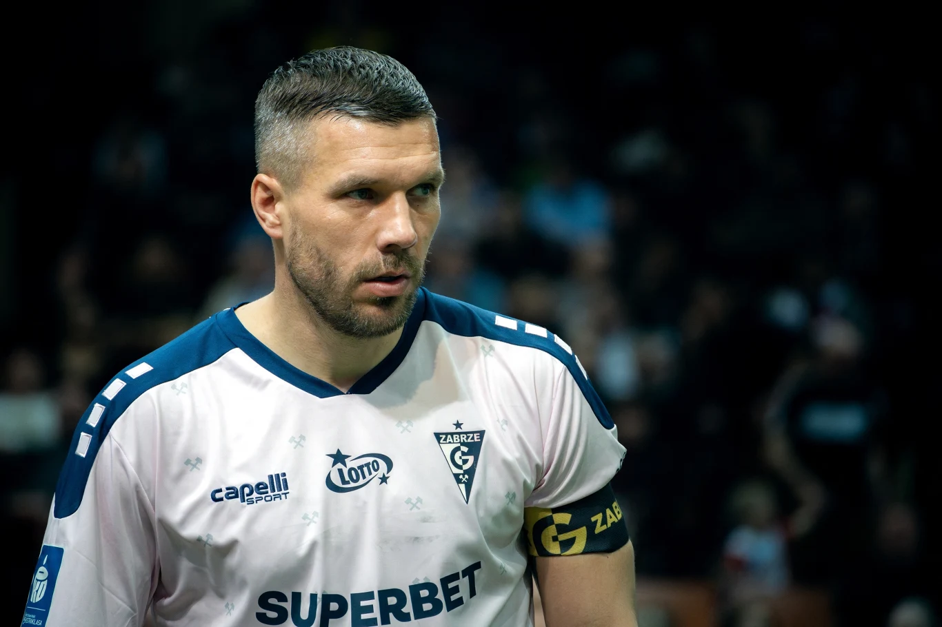 Lukas Podolski Piłkarz w biało-niebieskiej koszulce z opaską kapitana na ramieniu, widoczne logo klubu Górnik Zabrze oraz sponsorzy, w tle rozmyta publiczność.
