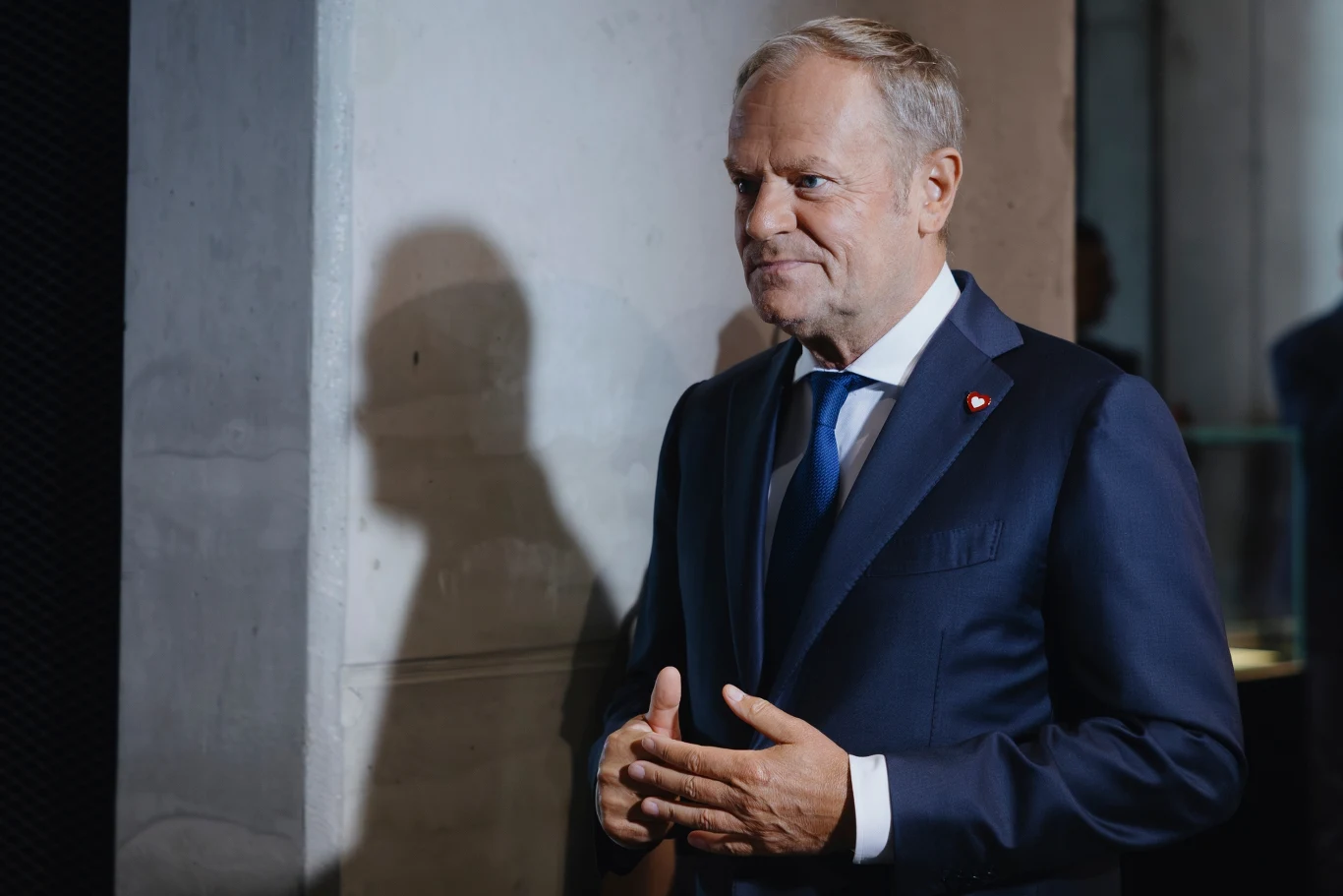 Premier Donald Tusk Mężczyzna w garniturze i niebieskim krawacie stoi przy jasnej ścianie, patrzy z lekkim uśmiechem na twarzy, w tle odbija się jego cień.