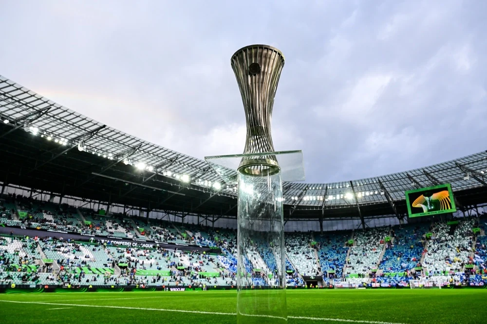 Trofeum Ligi Konfernecji Puchar konferencji ustawiony na środku boiska piłkarskiego na stadionie z zadaszonymi trybunami, na których widać tłum kibiców oraz elementy oprawy wizualnej.
