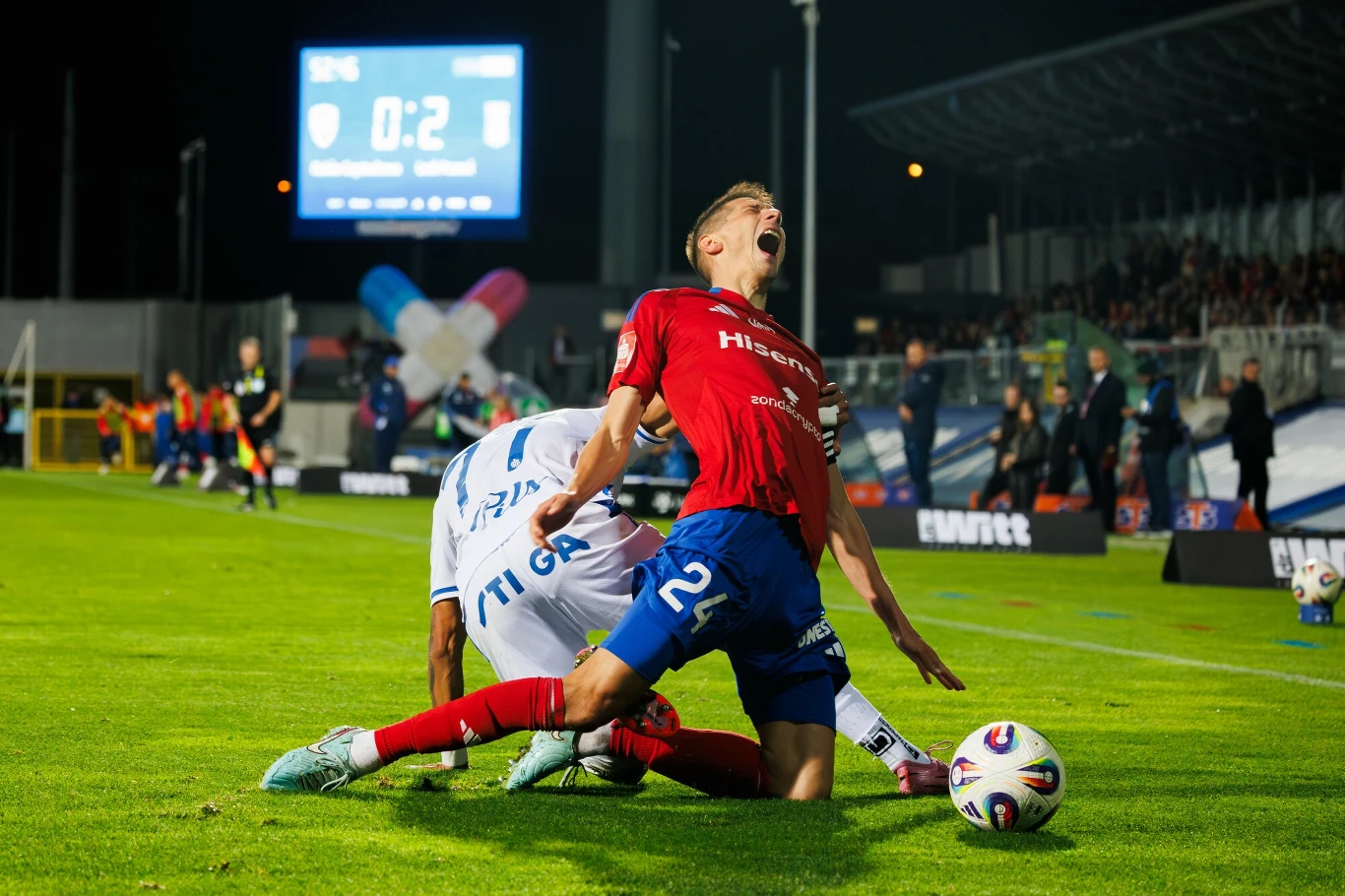 Zawodnik w czerwonym stroju podczas dynamicznej akcji piłkarskiej na tle boiska nocą, upada po kontakcie z rywalem ubranym na biało, a jego twarz wyraża ból i emocje, w tle widać fragment trybun oraz jasny wynik meczu na dużym ekranie.