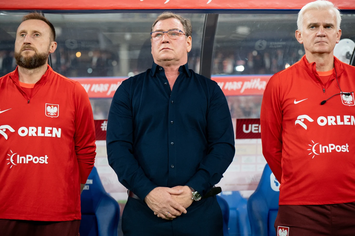 Trzech mężczyzn stojących obok siebie na tle stadionu piłkarskiego, dwóch z nich ubranych w czerwone dresy z logotypem Polskiego Związku Piłki Nożnej oraz sponsorów, środkowy w ciemnej koszuli, skupieni i poważni.